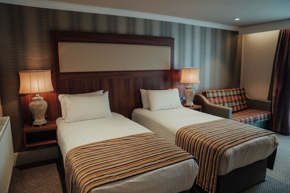 Quarto de hotel com duas camas, uma mesa de cabeceira com luminárias, uma cadeira e uma cabeceira grande.