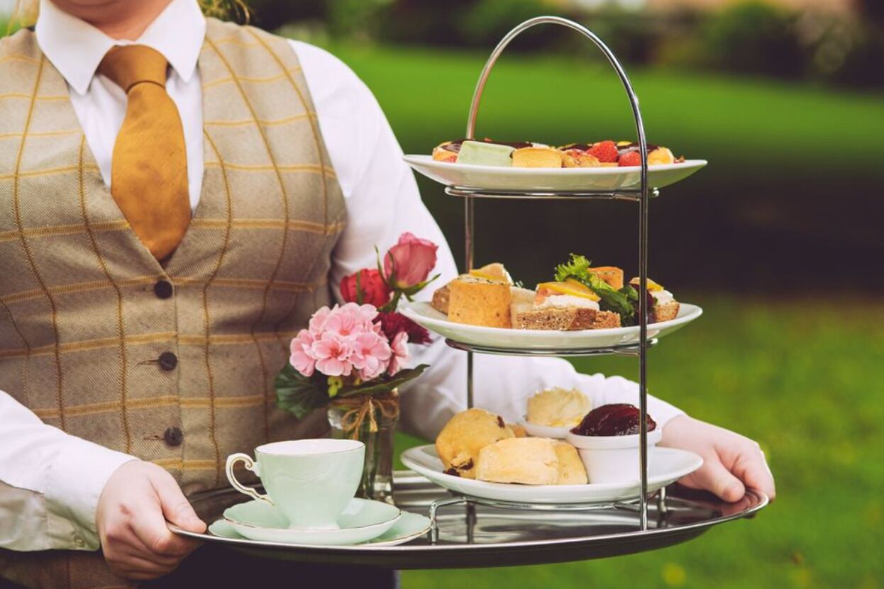 A person holding a tray with a three-tiered serving of pastries and sandwiches, a cup, and a vase with flowers.