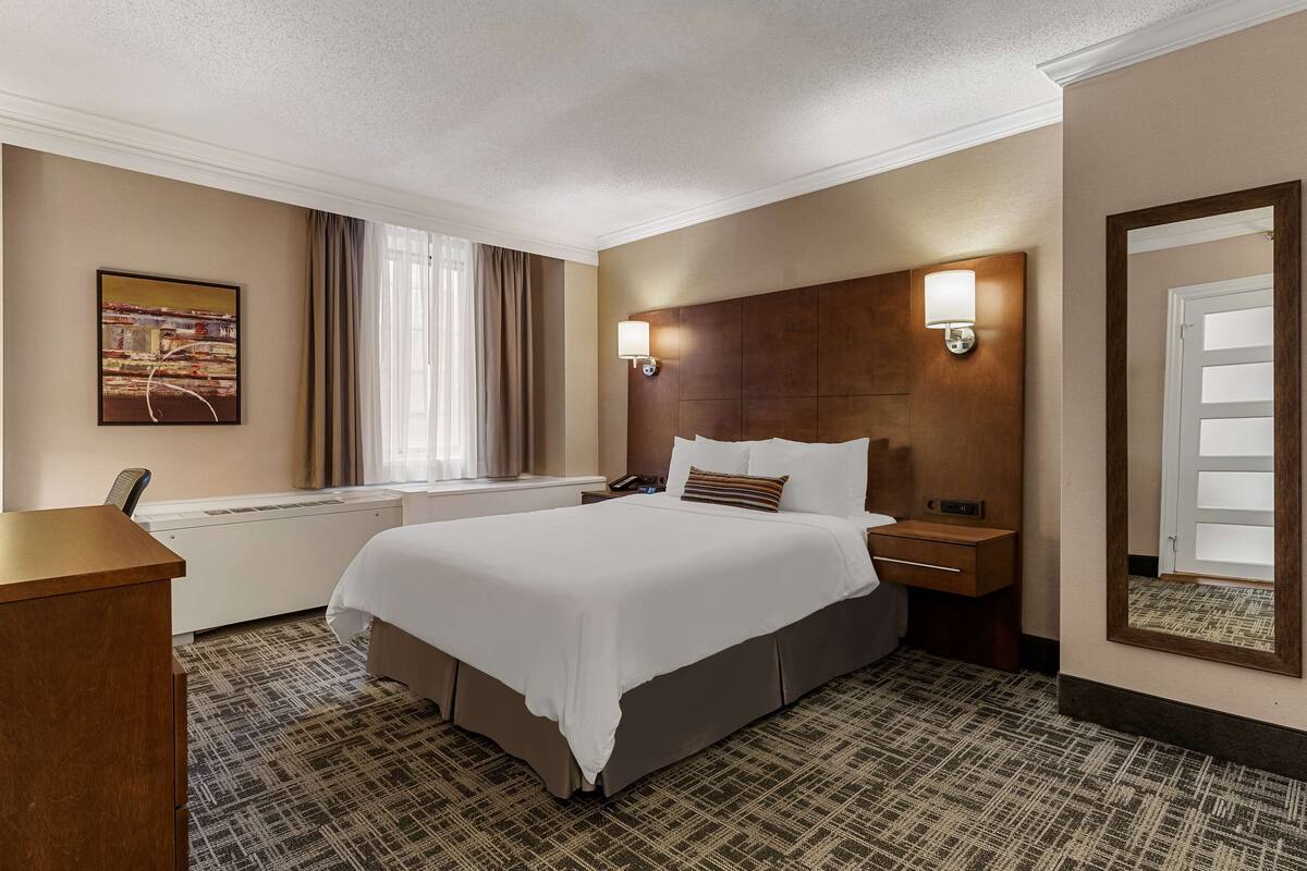 Habitación de hotel con una cama grande, un escritorio, una mesita de noche, una lámpara de pared, una ventana con cortinas y un cuadro en la pared.