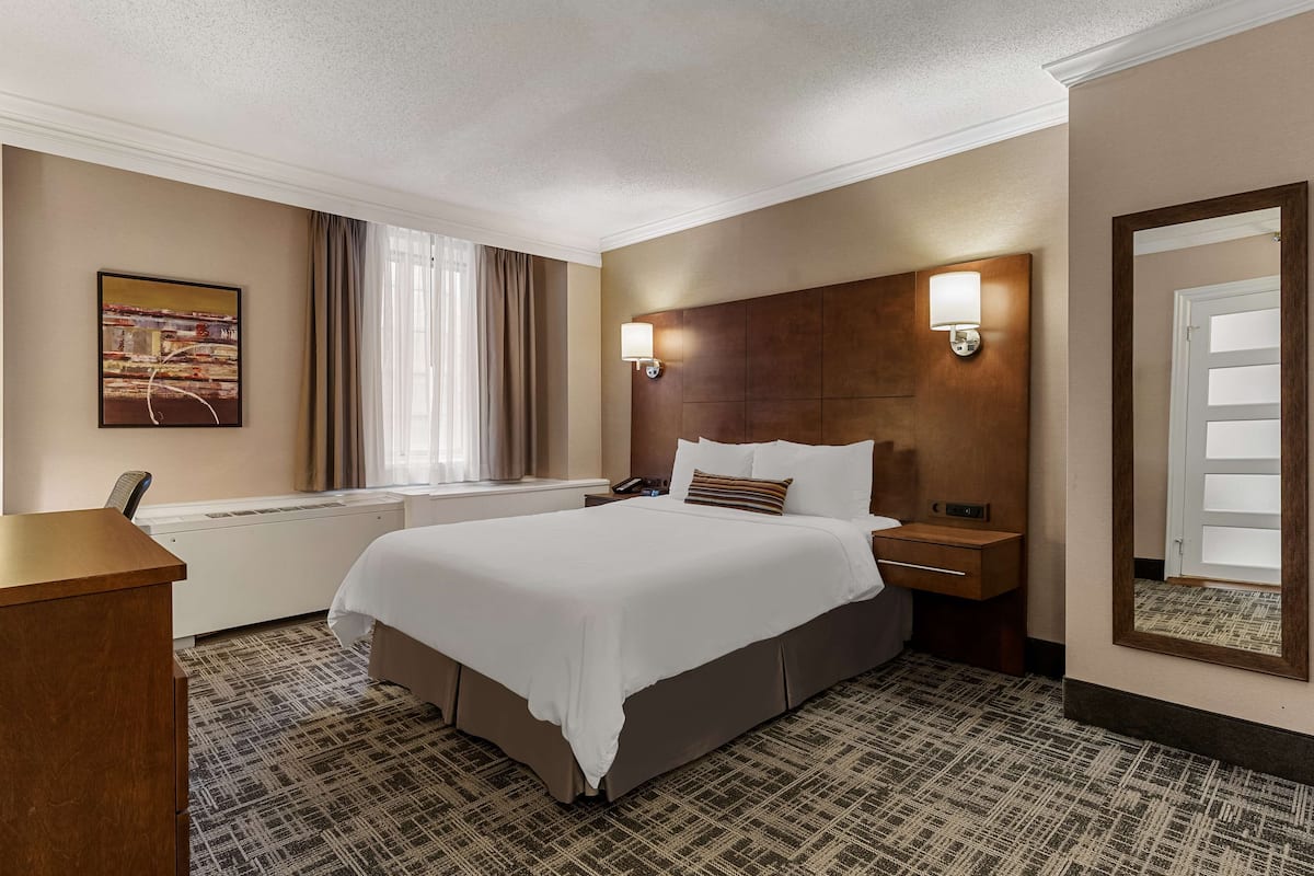 Quarto de hotel com uma cama grande, uma mesa de escritório, uma mesa de cabeceira, uma luminária na parede, uma janela com cortinas e um quadro na parede.