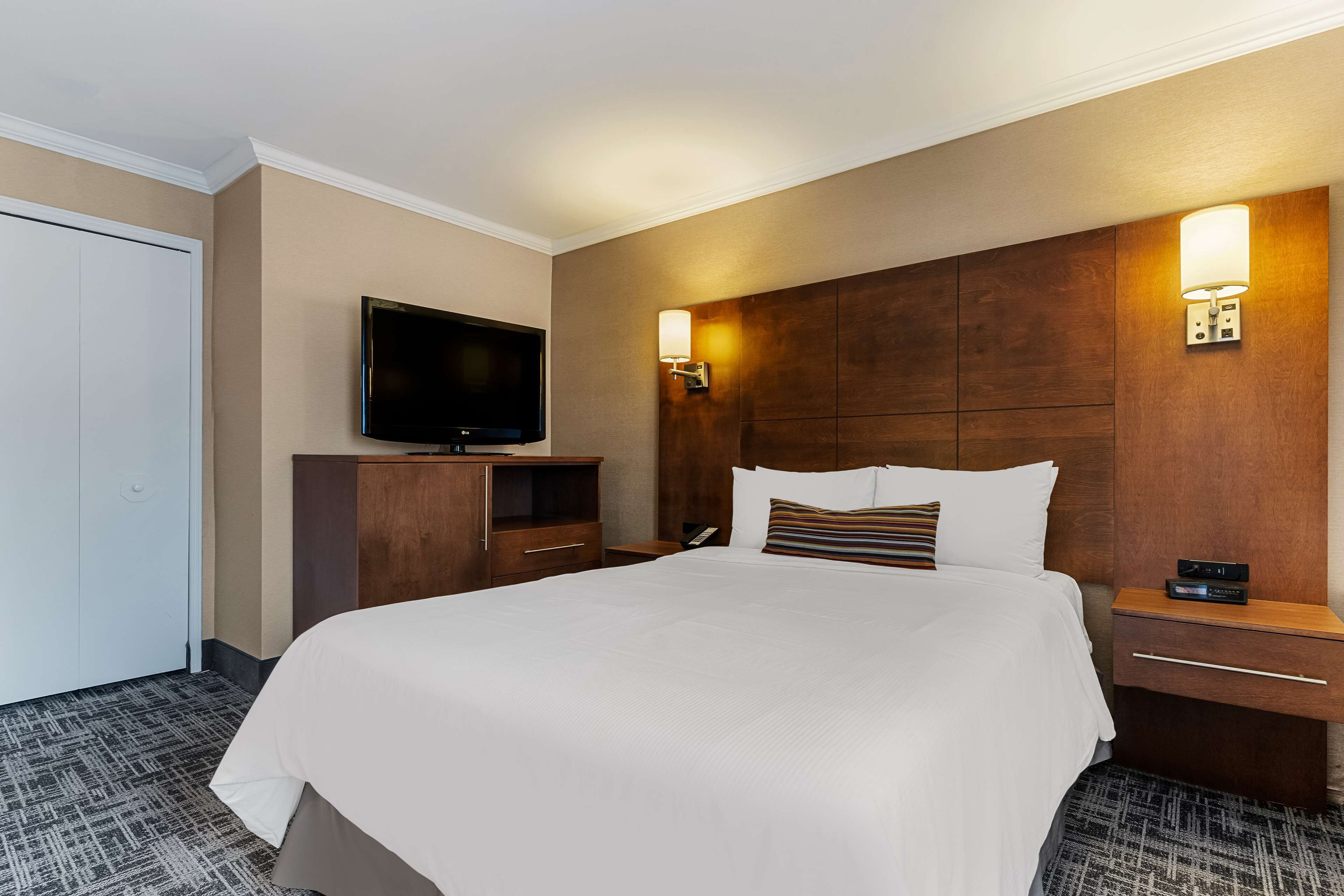 A hotel room with a large bed, a flat-screen TV, wooden headboard, bedside tables, and a wardrobe.
