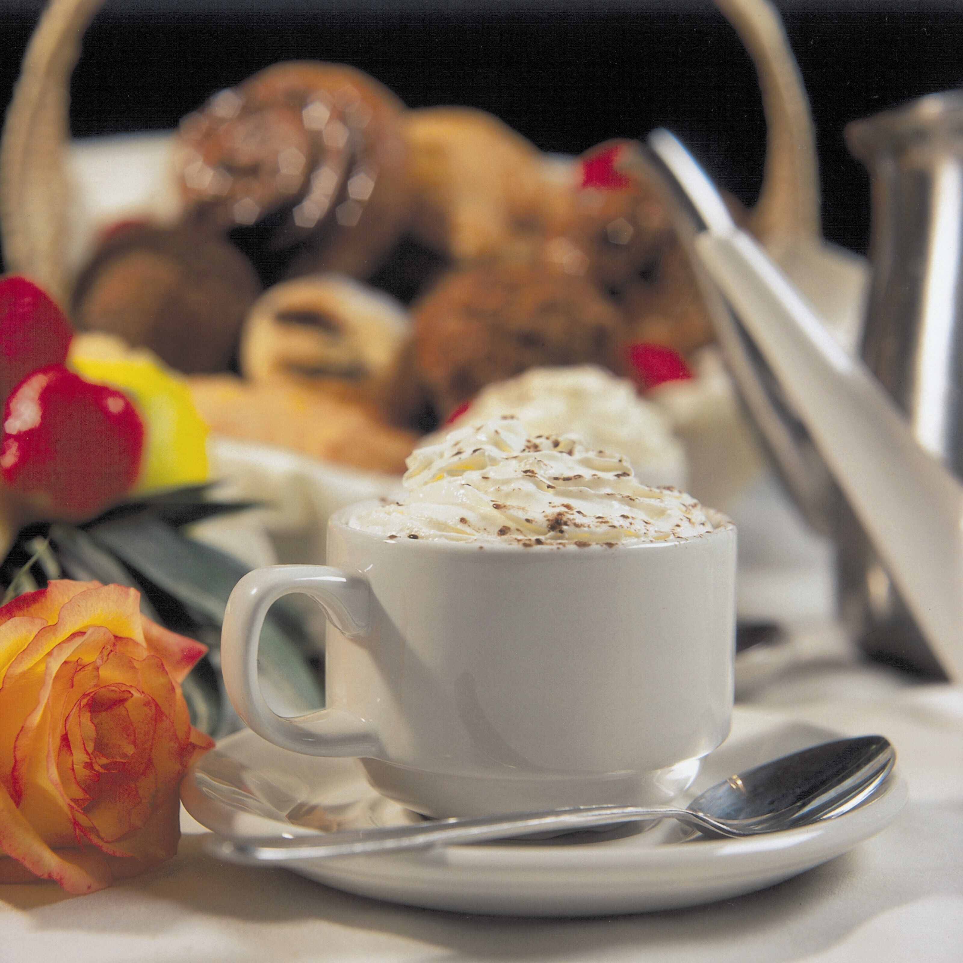 A white cup of coffee with whipped cream on a saucer, a spoon, and a basket of pastries in the background.