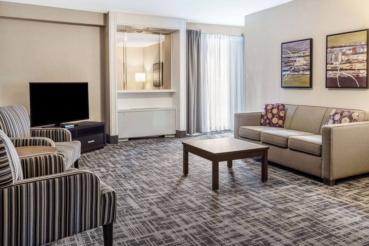 Una habitación de hotel moderna con un sofá a rayas, un sofá beige con cojines estampados, un televisor de pantalla plana, una mesita y un ventanal con cortinas.