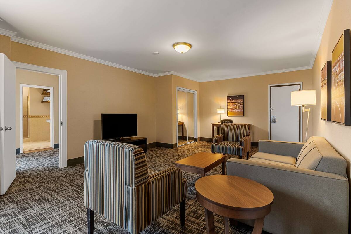 Habitación de hotel con sofá, sillones, televisor, lámpara y puerta que da a un baño.