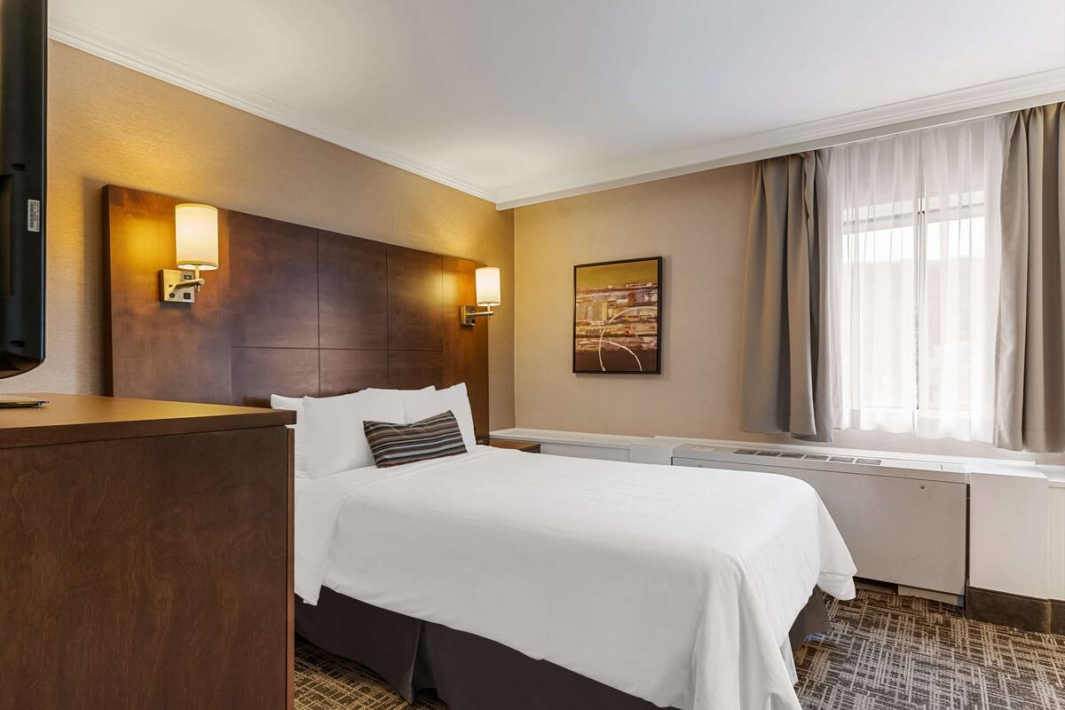Habitación de hotel con una cama grande, cabecera de madera, dos lámparas de pared, televisor de pantalla plana, ventana con cortinas e imagen enmarcadas en la pared.
