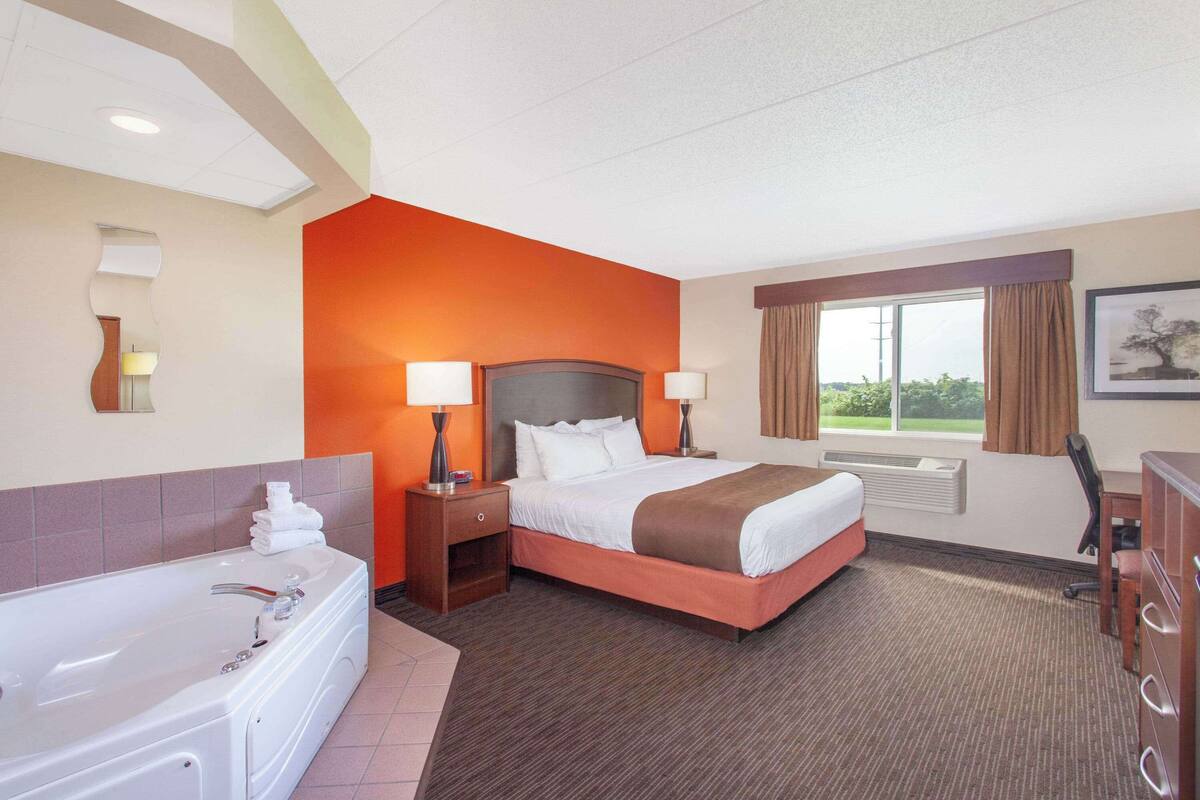 A hotel room with a large bed, a separate bathroom with a bathtub, a desk, and a window with curtains.
