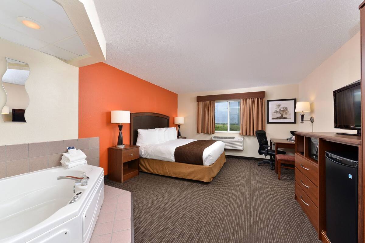 A hotel room with a large bed, a desk, a chair, a television, a bathtub, and a window with curtains.