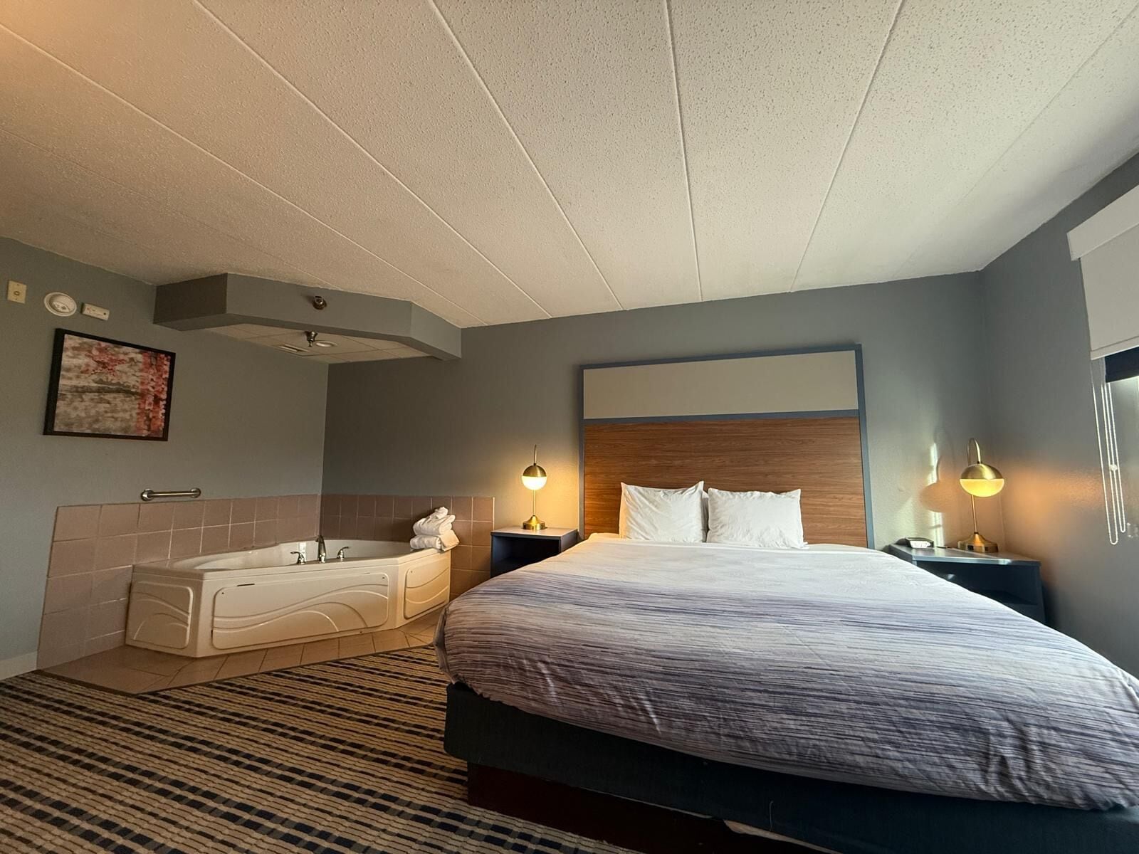 Ein Hotelzimmer mit Bett, Badewanne, zwei Nachttischlampen, einem Kopfteil und einem gemusterten Teppich.