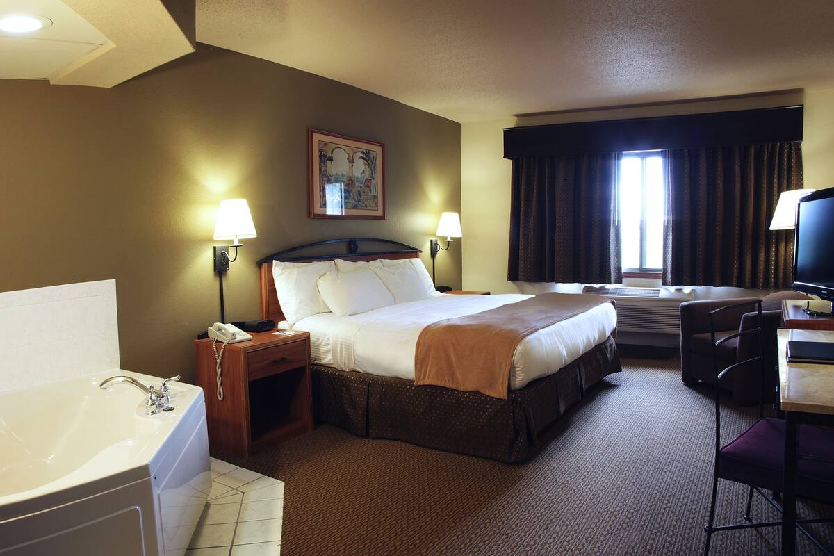 Habitación de hotel con una cama grande, un escritorio con silla, una lámpara de escritorio, un teléfono, un cuadro en la pared y una ventana con cortinas.