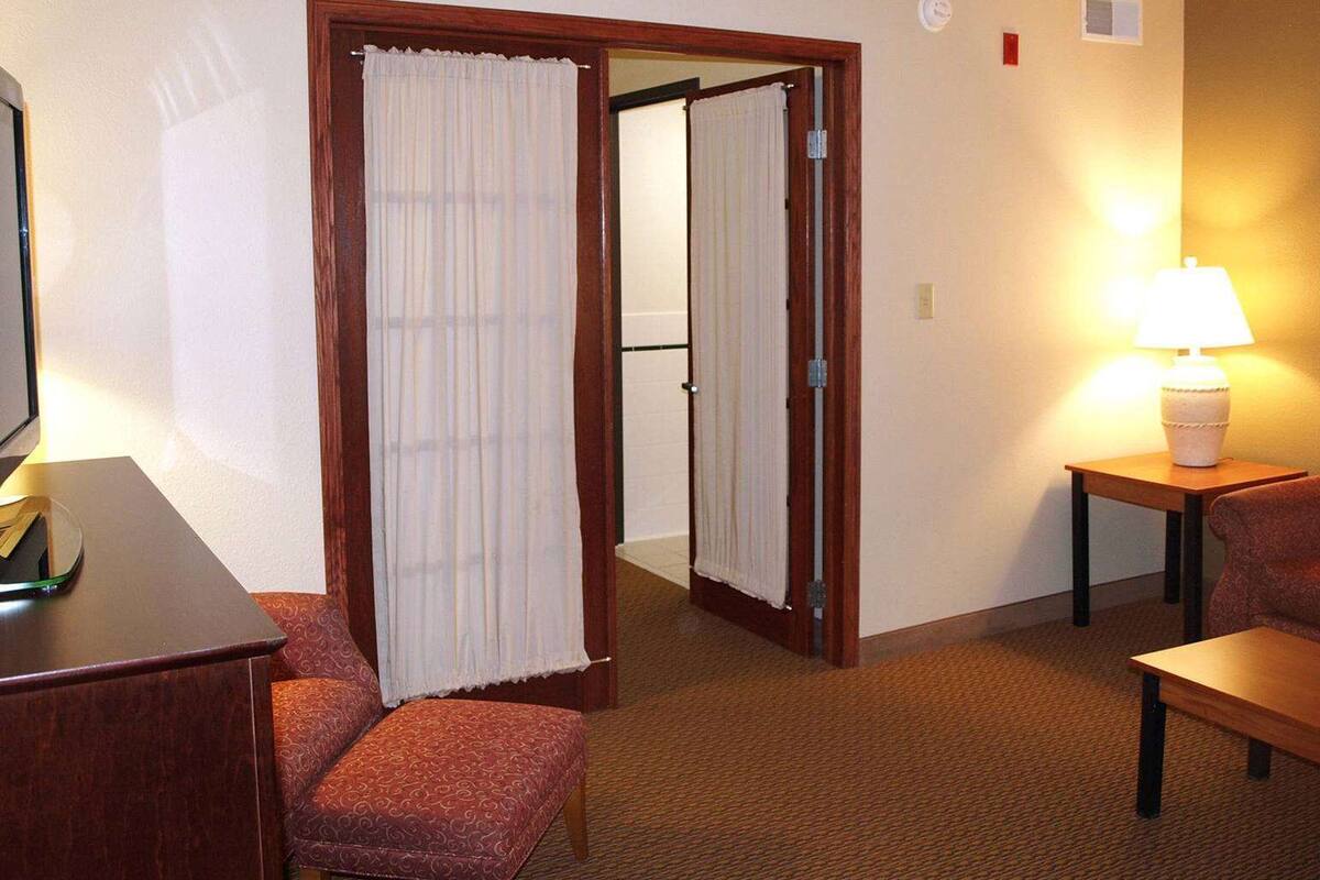 Una habitación de hotel con cama, escritorio, silla, lámpara y puerta que da a otra estancia.