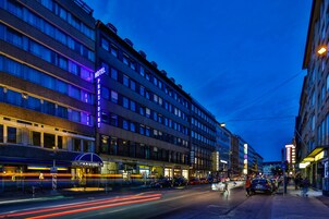 Un hotel con diversi piani, un cartello ben visibile e una strada trafficata con auto e pedoni.