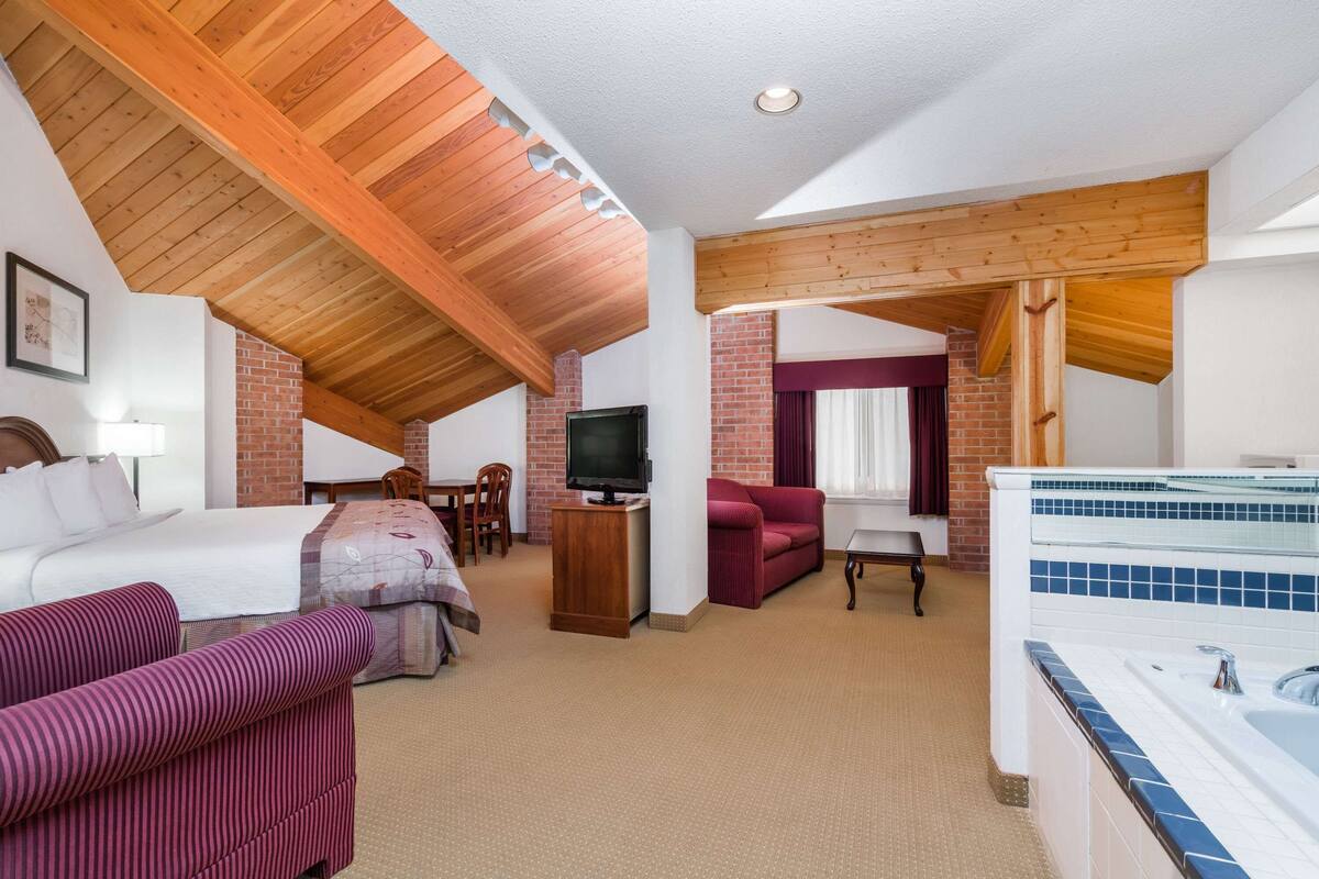 Une chambre d’hôtel avec un plafond haut, des poutres apparentes, une cheminée en briques, une télévision, un canapé, un lit, une petite table et une baignoire.