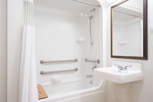 Un baño con bañera, barras de apoyo, ducha, lavamanos y espejo.