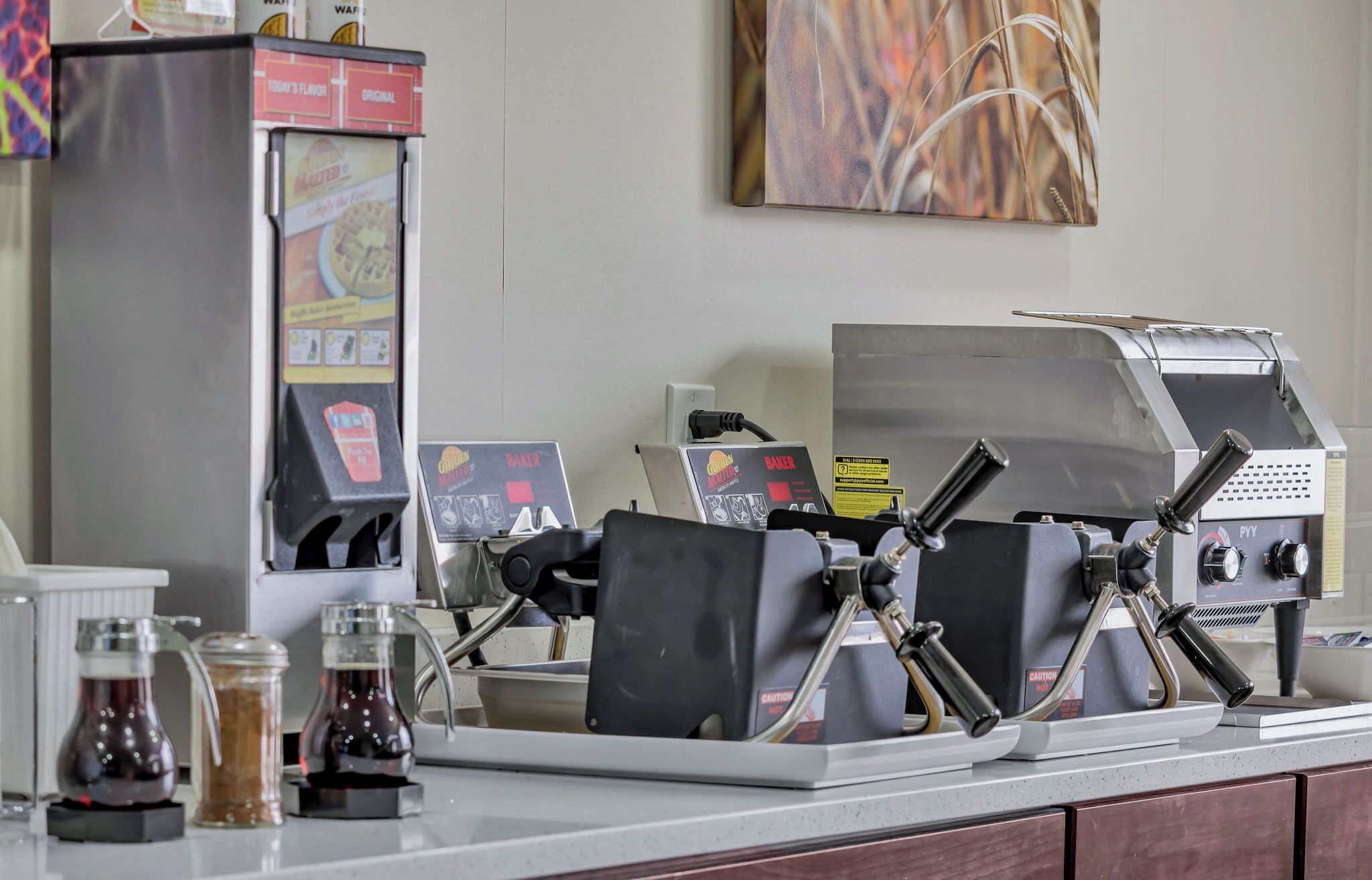 Une machine à café et un appareil à gaufres sont posés sur un comptoir, à côté d’un distributeur de café et de pots contenant divers condiments.