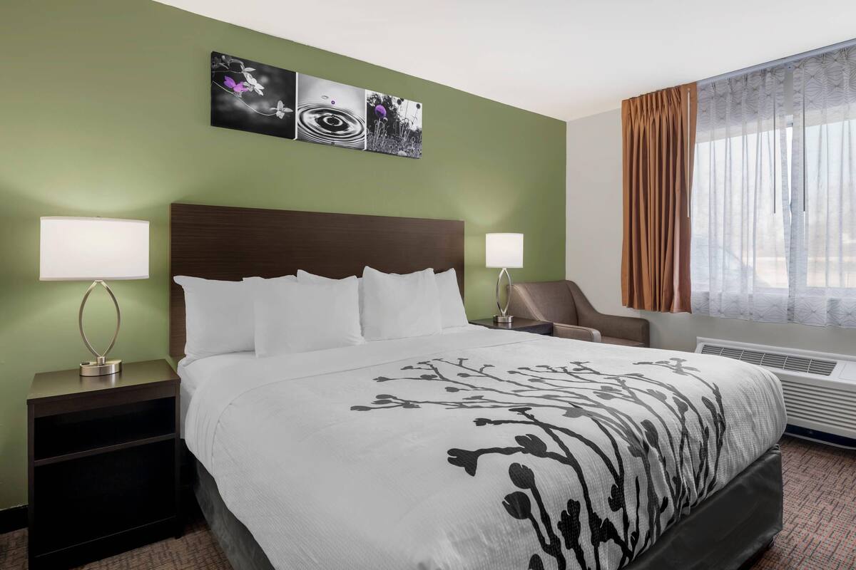 Habitación de hotel con una cama grande, dos mesitas de noche con lámparas, una silla, un ventanal con cortinas y decoración en la pared.