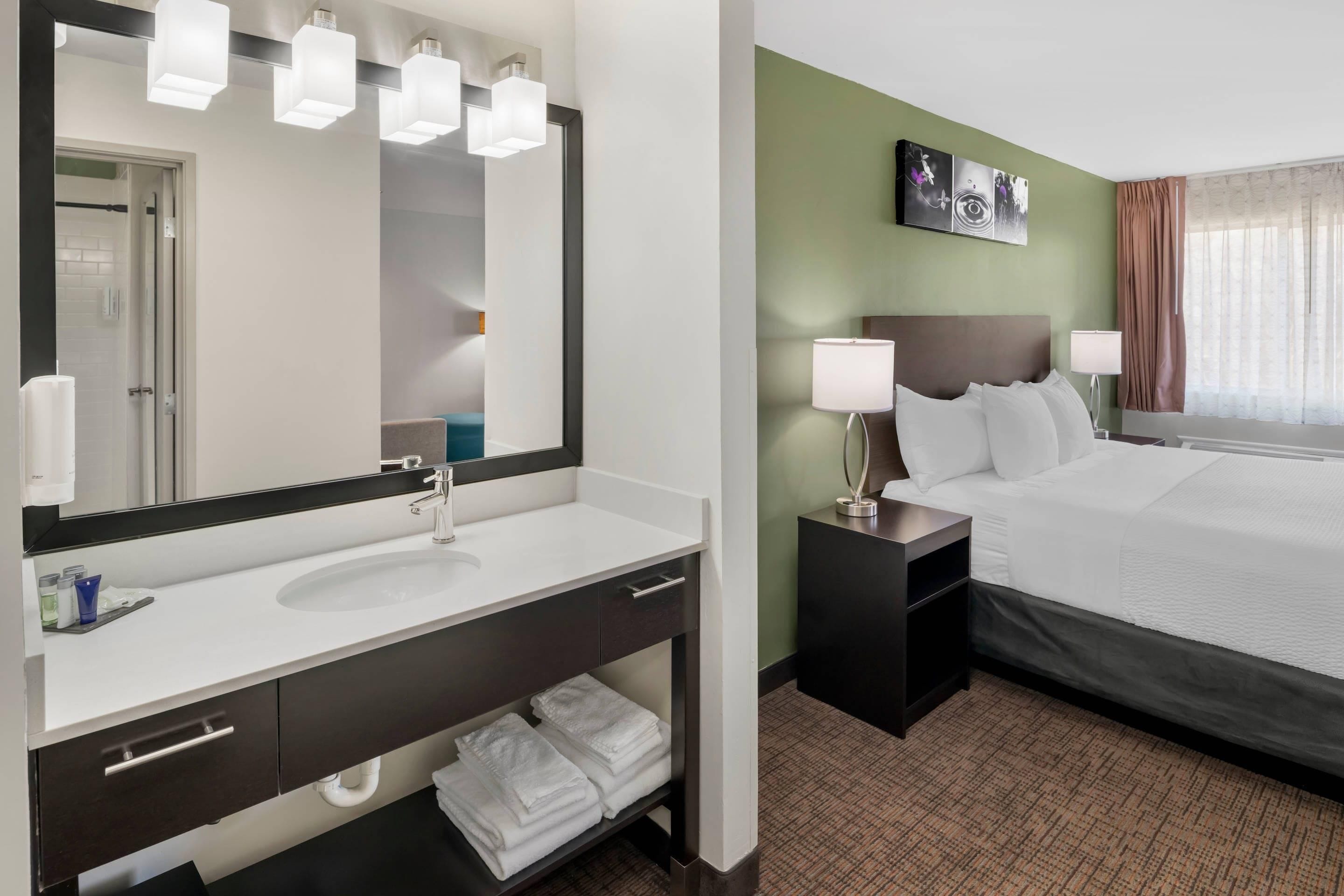 A hotel room with a large mirror, a white sink, a bed, a nightstand, and a wall-mounted artwork.