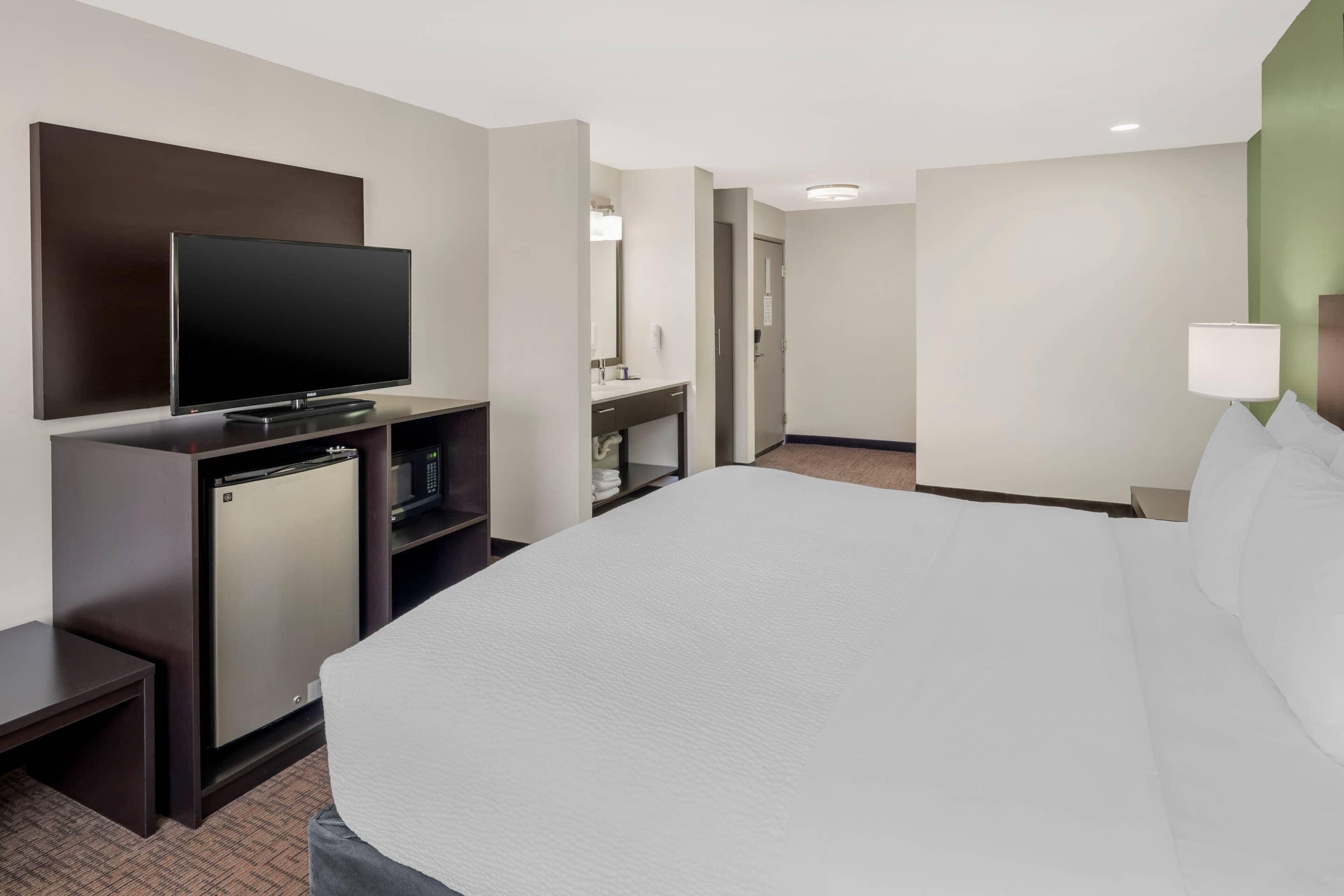 A hotel room with a bed, a TV, a mini-fridge, a desk, a lamp, and a bathroom door.