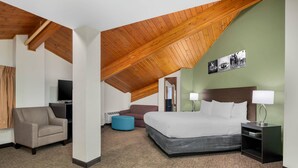 A hotel room with a high wooden ceiling, a bed with white linens, a blue ottoman, a brown armchair, and a television.