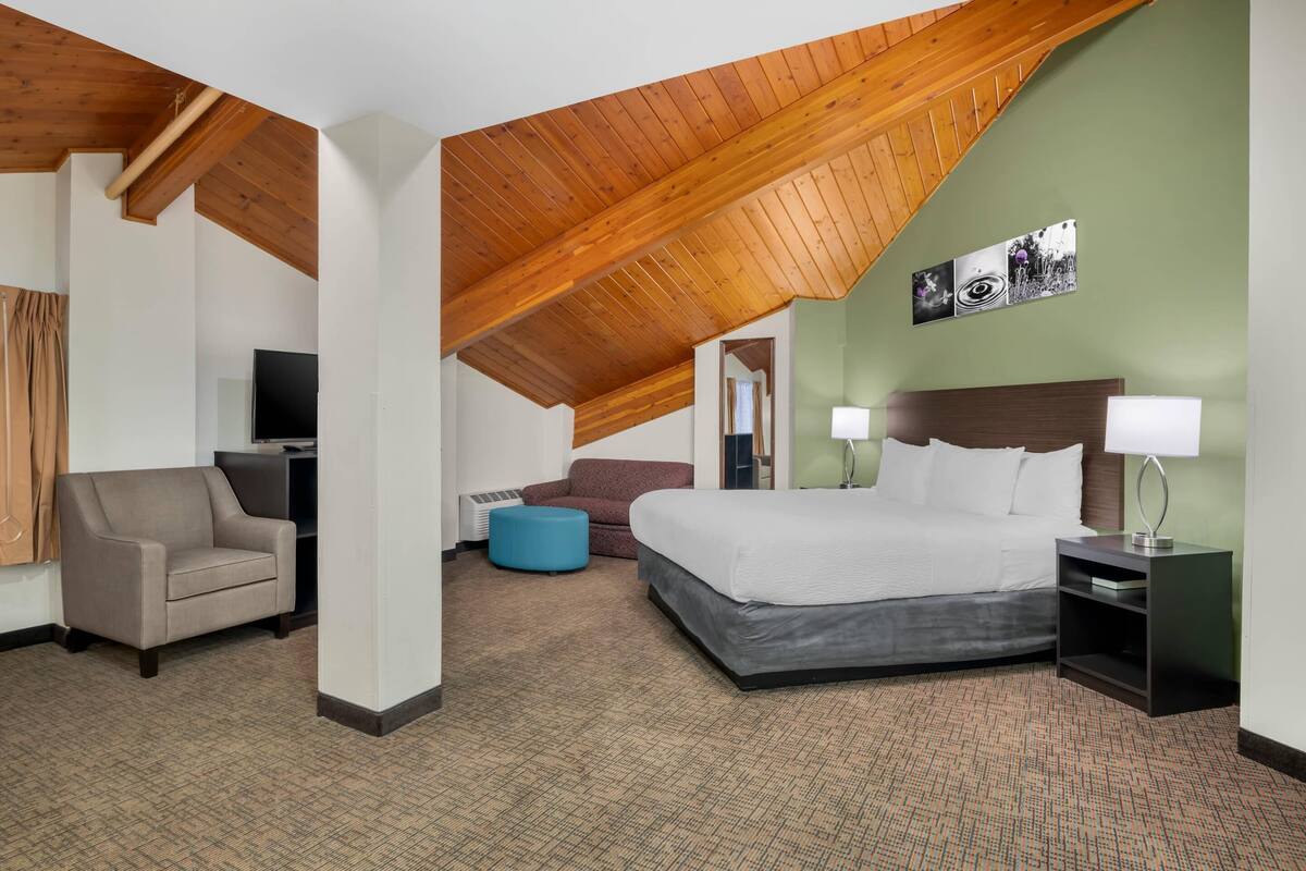 Habitación de hotel con un alto techo de madera, una cama con ropa blanca, un reposapiés azul, un sillón marrón y un televisor.