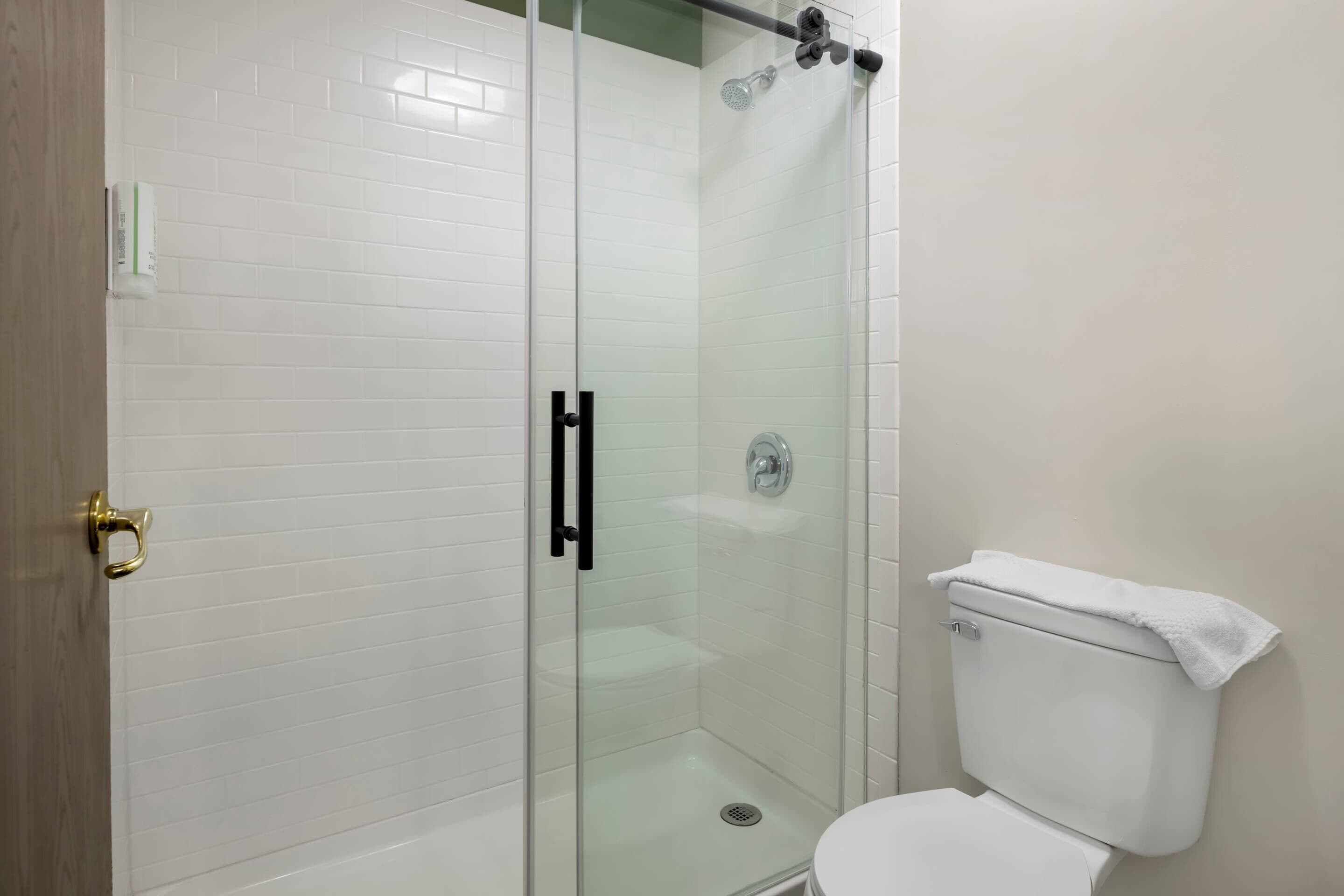A glass-enclosed shower with a white tiled wall, a white toilet, and a gold door handle.