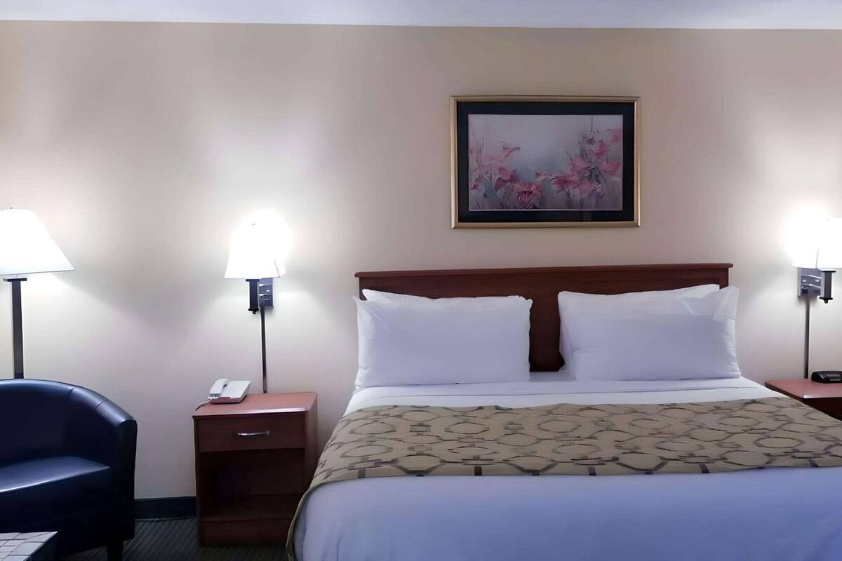 Habitación de hotel con cama, dos lámparas de noche, una silla, mesita de noche, teléfono y un cuadro en la pared.