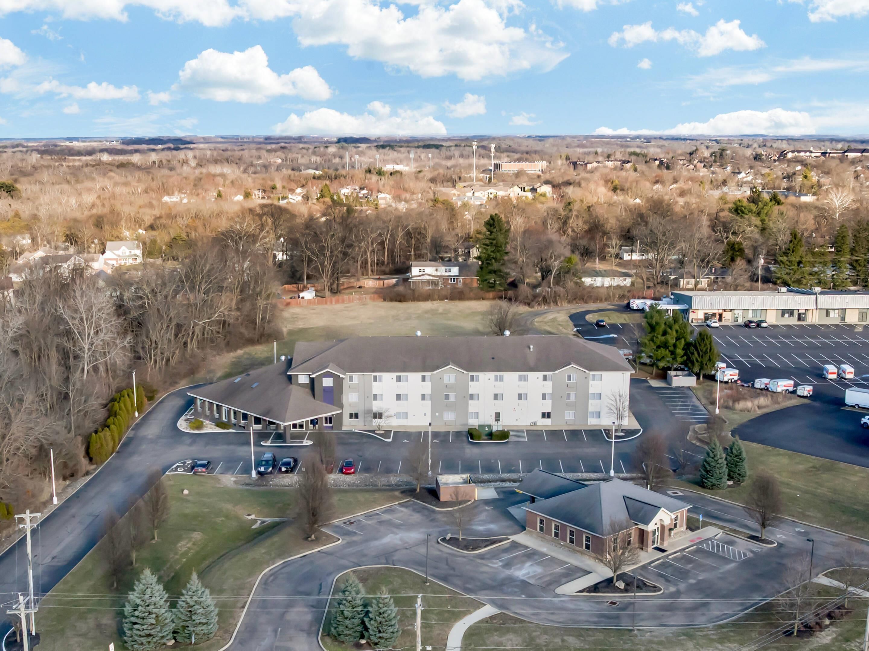 An aerial view of a multi-story hotel with a parking lot, surrounded by trees and a distant view of a town.