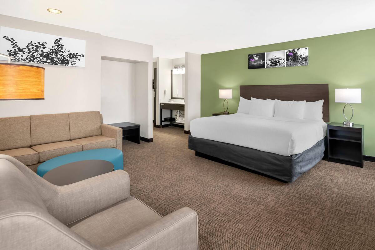 Habitación de hotel con cama, sofá, escritorio y baño visible a través de una puerta abierta.