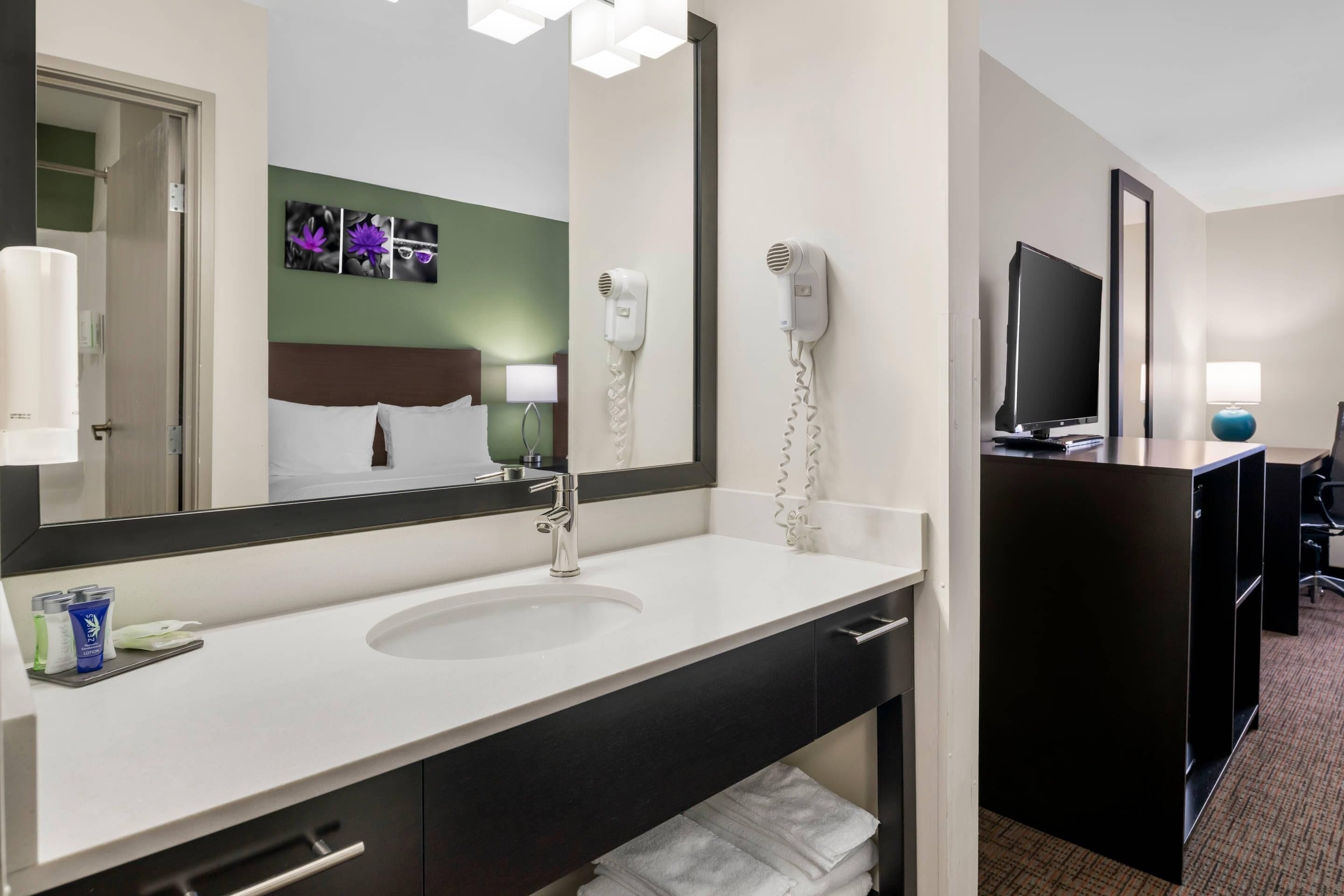 Baño de hotel con lavamanos, espejo, secador de pelo y televisor en la habitación contigua.