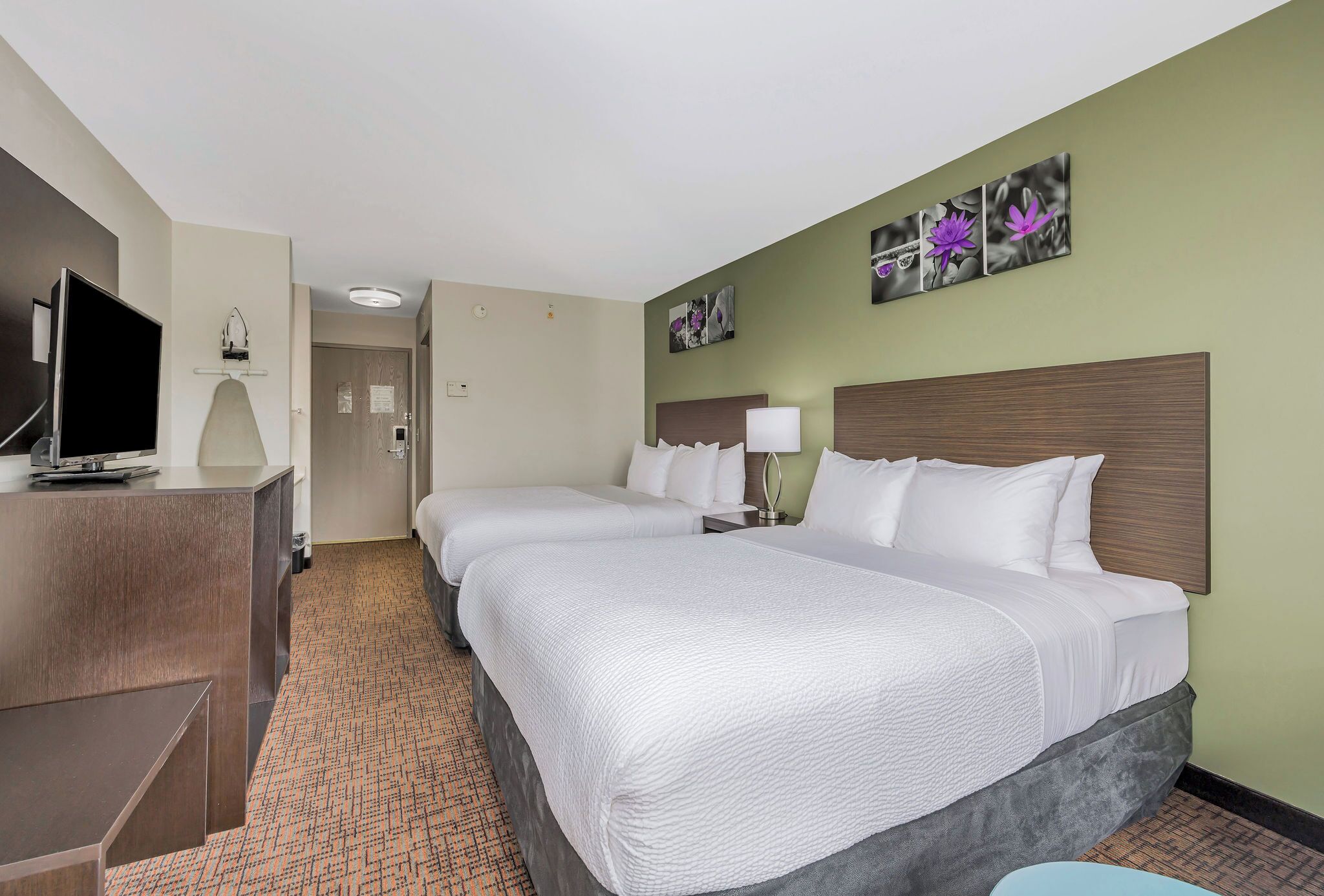Quarto de hotel com duas camas, uma mesa de trabalho com televisão de tela plana, uma luminária e obras de arte na parede.