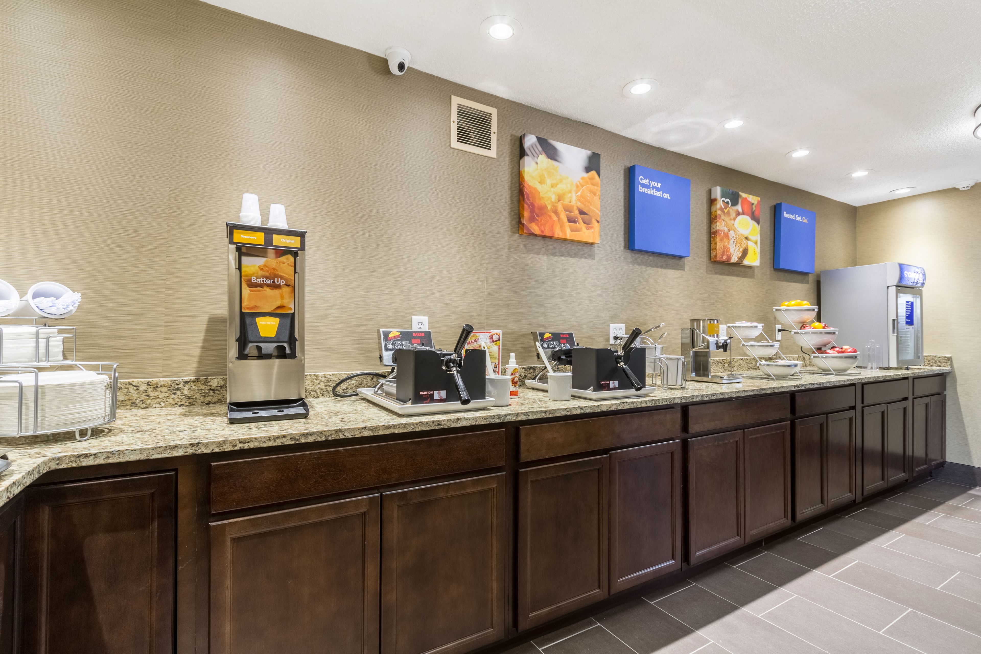 A self-service breakfast bar with coffee machines, a beverage dispenser, and a refrigerator.