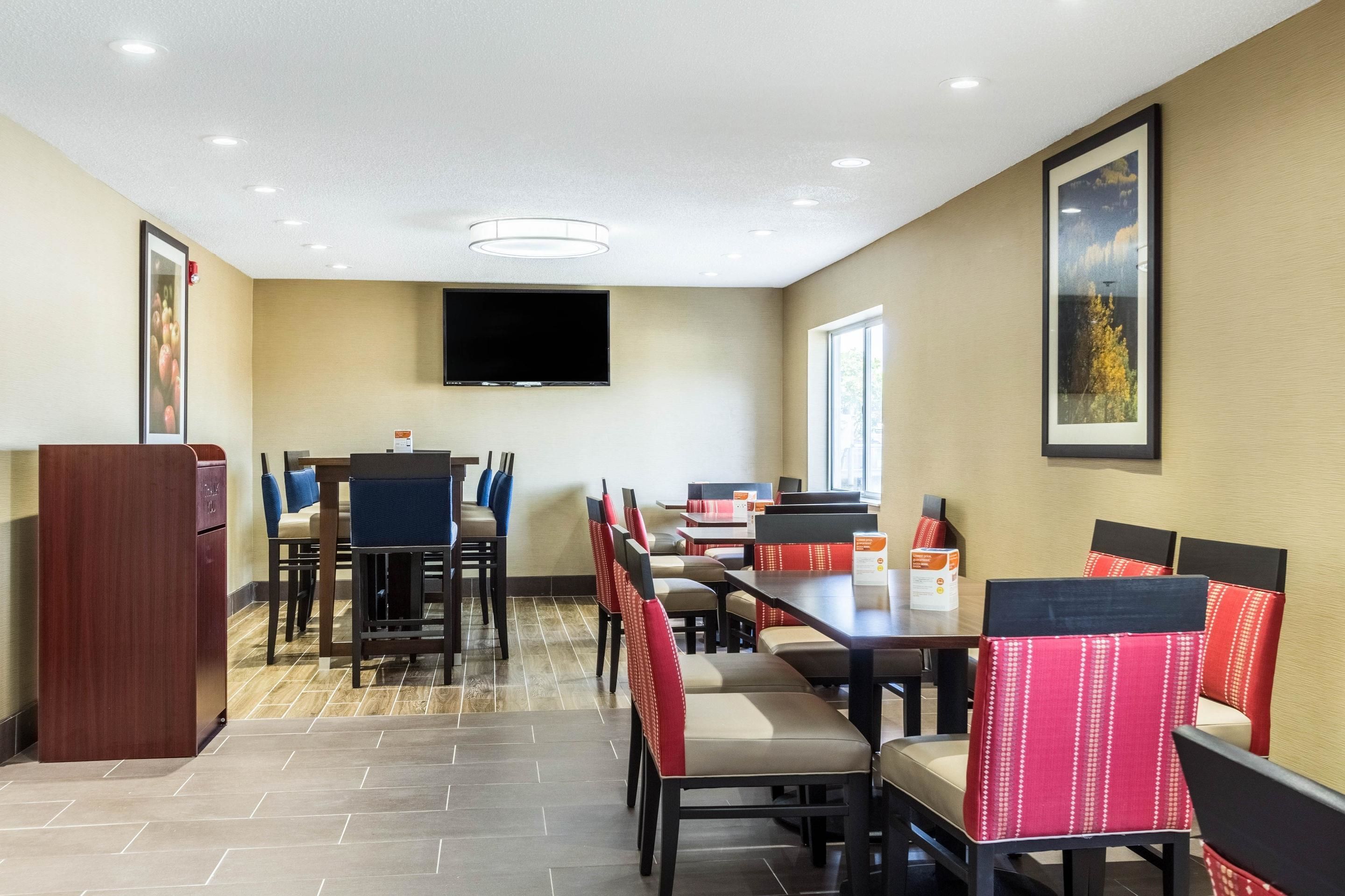 A hotel dining area with multiple tables and chairs, a flat-screen TV mounted on the wall, and framed pictures on the walls.