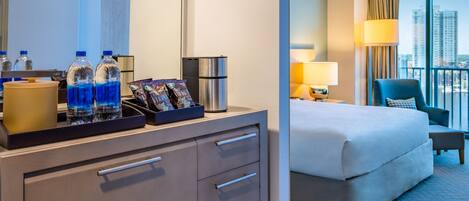 Habitación de hotel con cama, escritorio con cafetera, botellas de agua y un minibar.