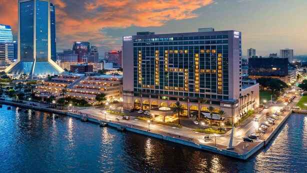 Un hôtel moderne, reconnaissable à son grand panneau « HYATT », situé au bord de l’eau et offrant une vue sur les gratte-ciel de la ville.