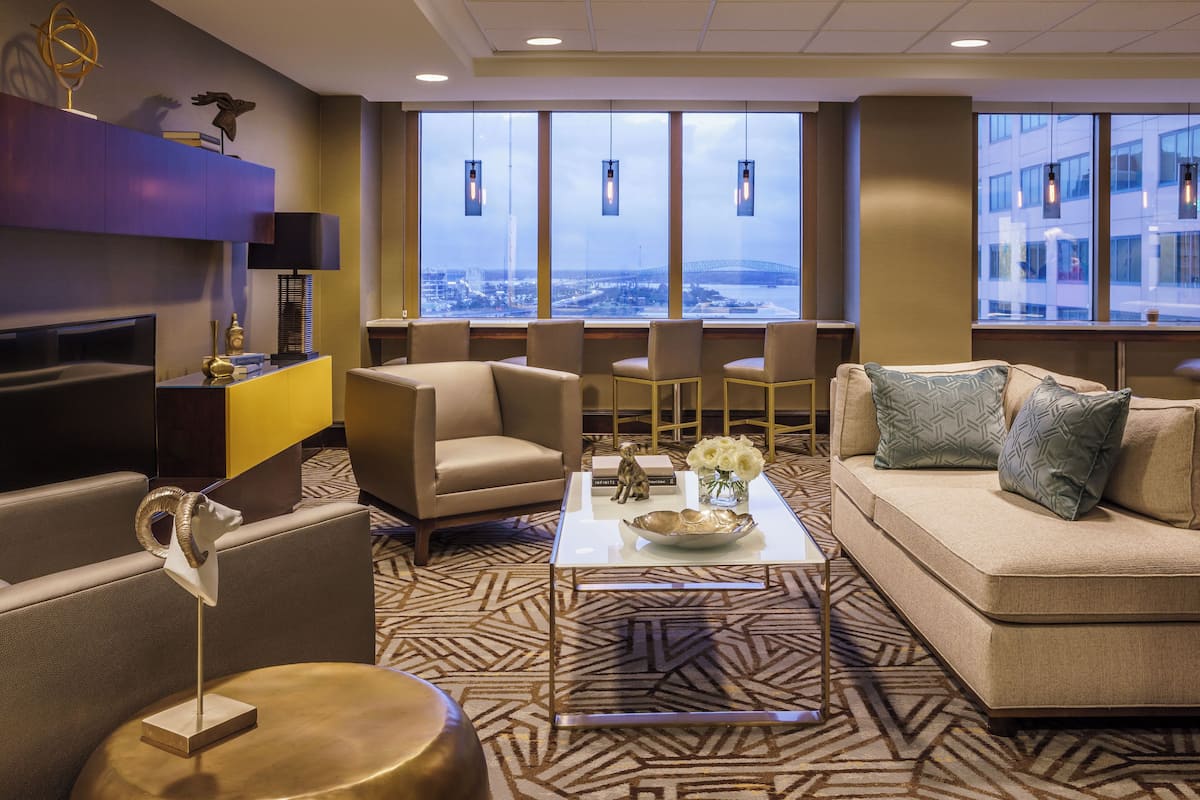 A modern hotel lobby with a sofa, armchairs, a coffee table, and a TV. Large windows offer a view of the city and a bridge.