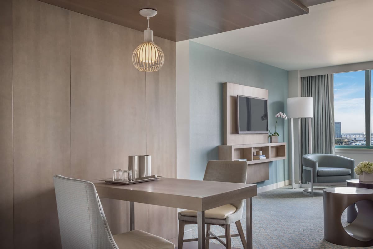A modern hotel room with a dining area, a sofa, a TV, and a view of the city.