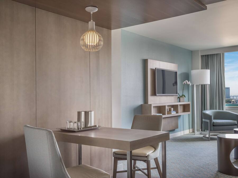 A modern hotel room with a dining area, a sofa, a TV, and a view of the city.