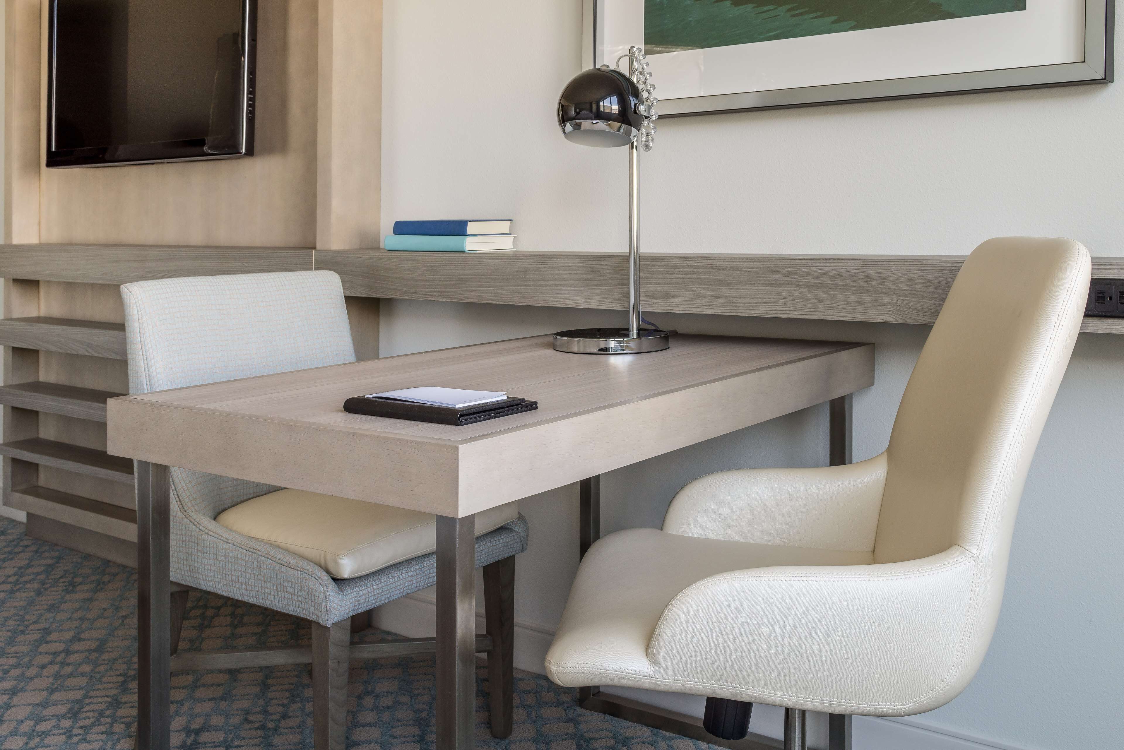 A modern hotel room with a desk, two chairs, a desk lamp, a flat-screen TV, and a framed picture on the wall.
