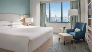 A hotel room with a large bed, a blue armchair, a small table with a lamp, and a view of a bridge.
