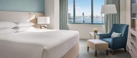 Habitación de hotel con una cama grande, un sillón azul, una mesita con lámpara y vistas a un puente.