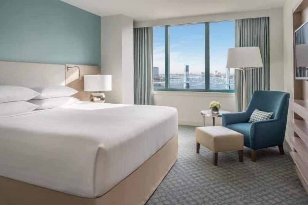 A hotel room with a large bed, a chair, a small table with a lamp, and a window with a city view.