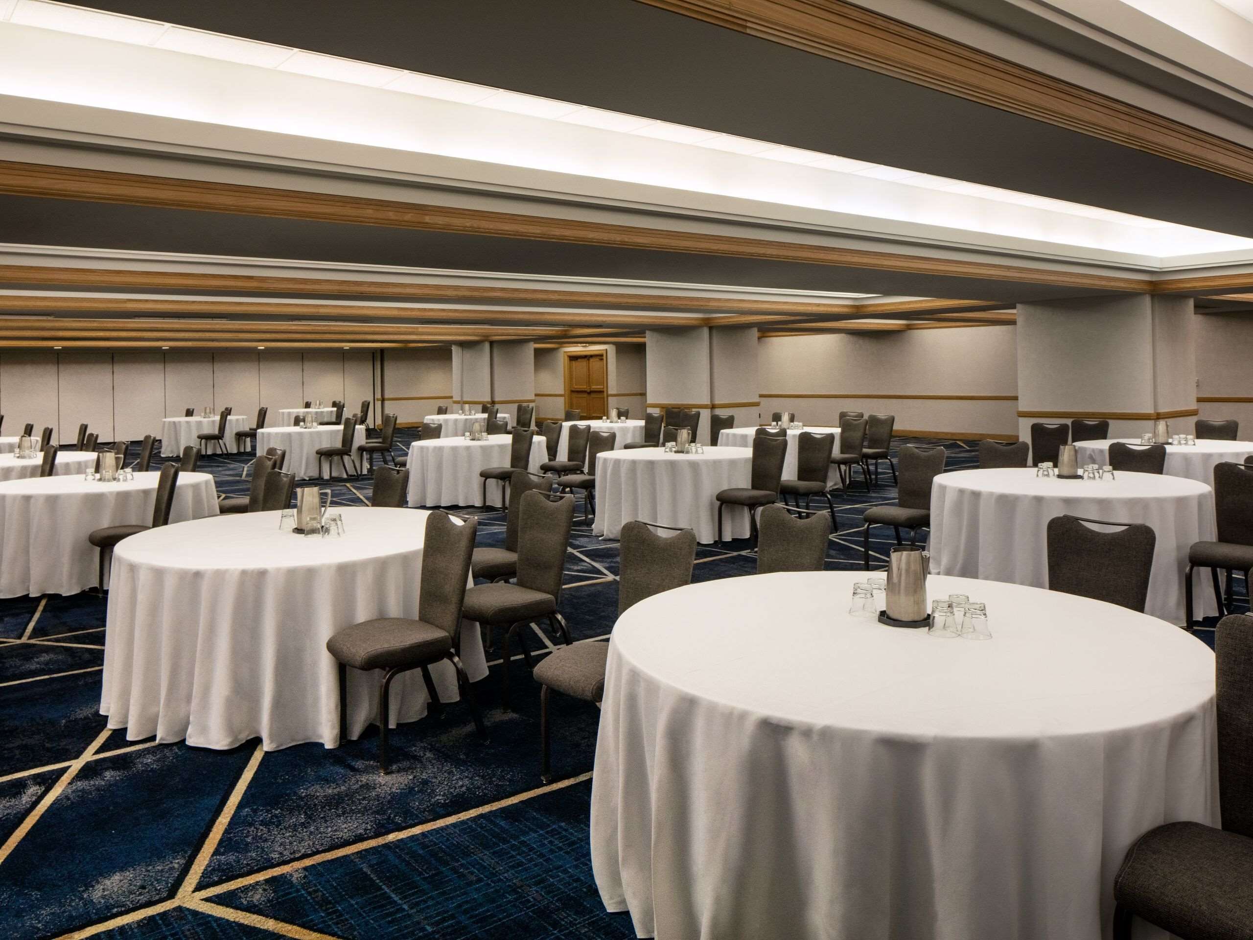 A large conference room with multiple round tables, each with a white tablecloth and a candle centerpiece. The room has a carpeted floor and a wooden ceiling with recessed lighting.