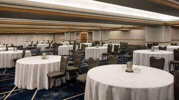 A large conference room with multiple round tables, each with a white tablecloth and a candle centerpiece. The room has a carpeted floor and a wooden ceiling with recessed lighting.