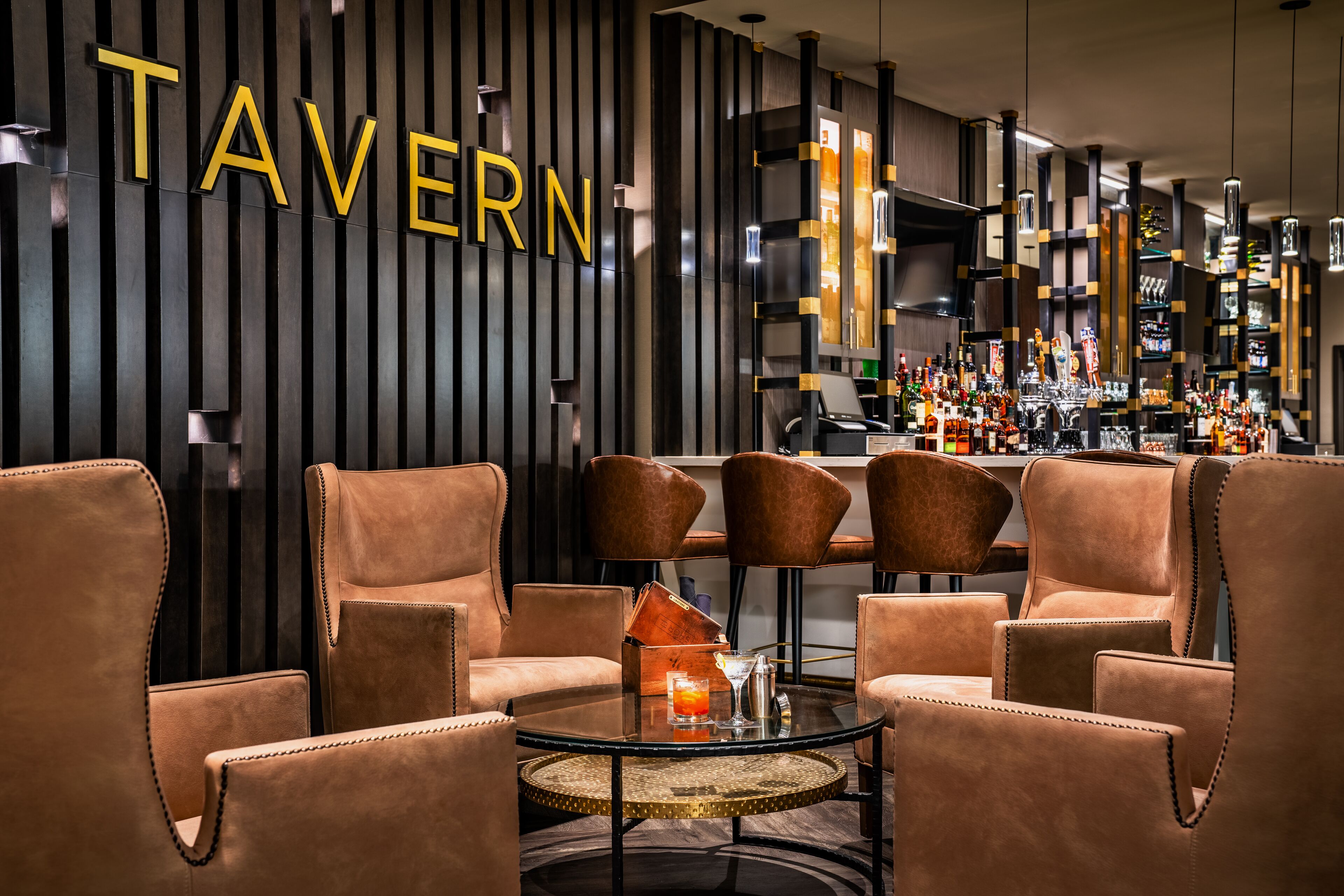 Un área de bar moderna con sillones de cuero, una barra y una pared con el letrero 'TAVERN'.