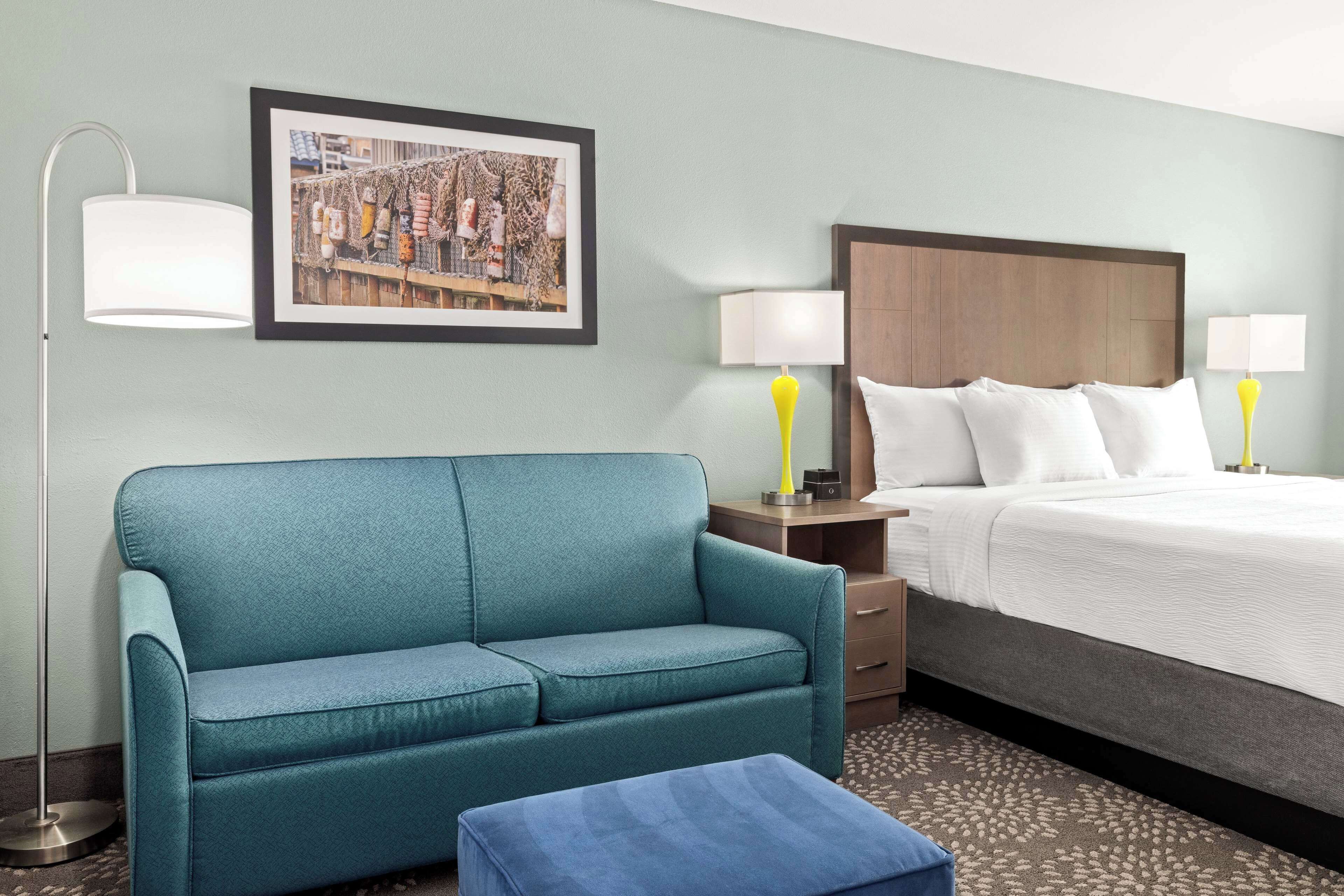 Una habitación de hotel con un sofá color turquesa, una cama con ropa blanca, dos lámparas de noche y un cuadro en la pared.