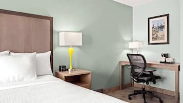 Habitación de hotel con cama, un escritorio con silla, lámpara, cafetera y un cuadro en la pared.