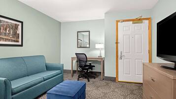 A room with a teal sofa, a blue ottoman, a desk with a chair, a television on a wooden cabinet, and a door with a mirror above it.