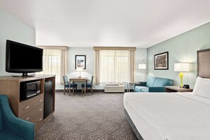 A hotel room with a bed, a desk with a chair, a television, a microwave, a mini-fridge, a sofa, a lamp, and a window with curtains.