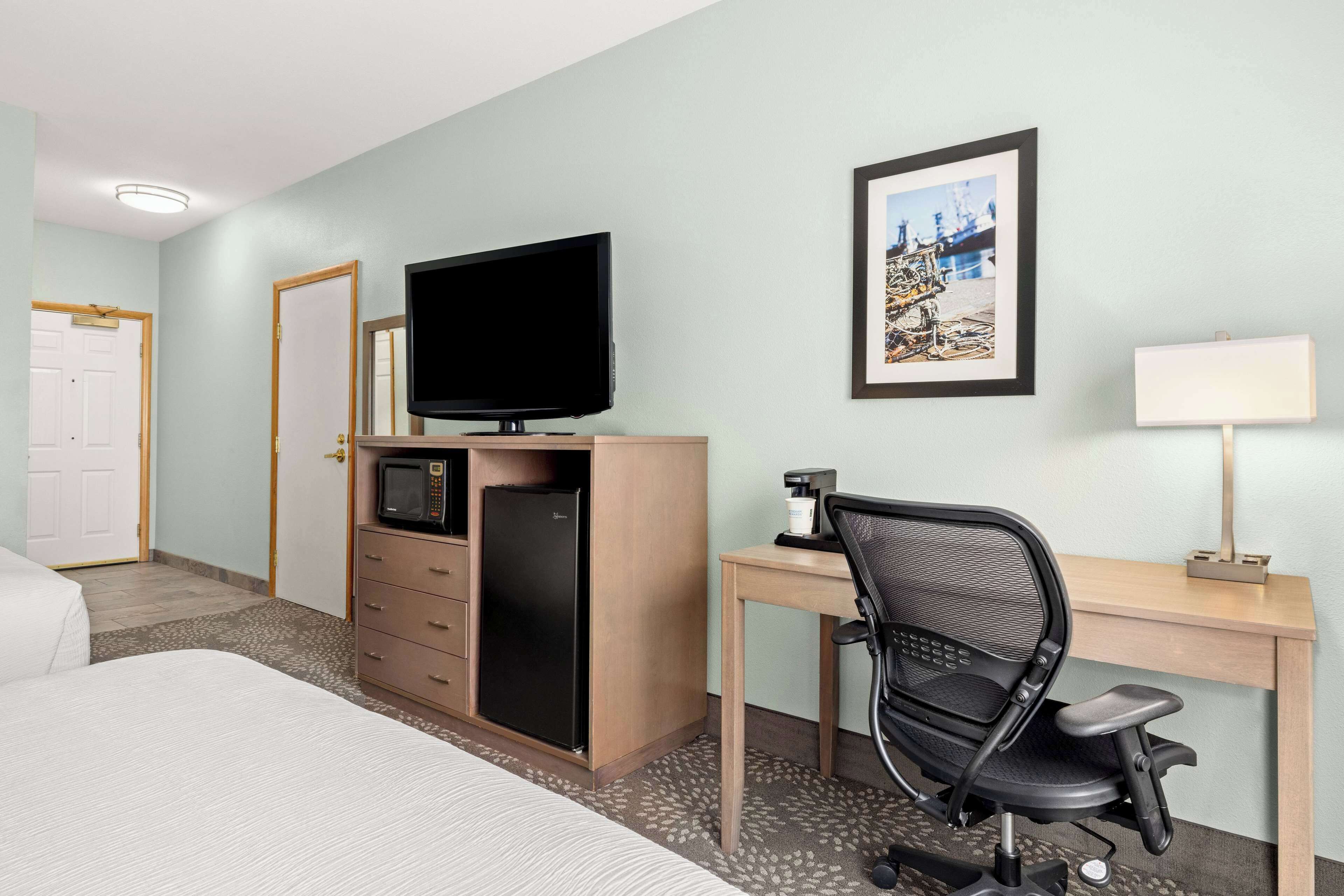 A hotel room with a bed, a desk with a chair, a television, a refrigerator, a microwave, a coffee maker, and a framed picture on the wall.