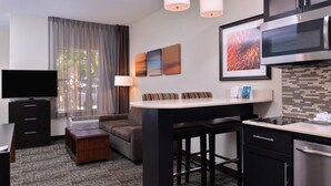 Une chambre d’hôtel moderne équipée d’une télévision à écran plat, d’un canapé, d’une petite table, d’un coin cuisine avec un four à micro-ondes et d’une fenêtre avec des rideaux.