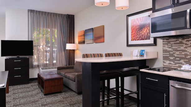 Ein kompaktes Hotelzimmer mit Küchenzeile, einem Flachbildfernseher, einem Sofa, einem Essbereich und einem großen Fenster mit Vorhängen.